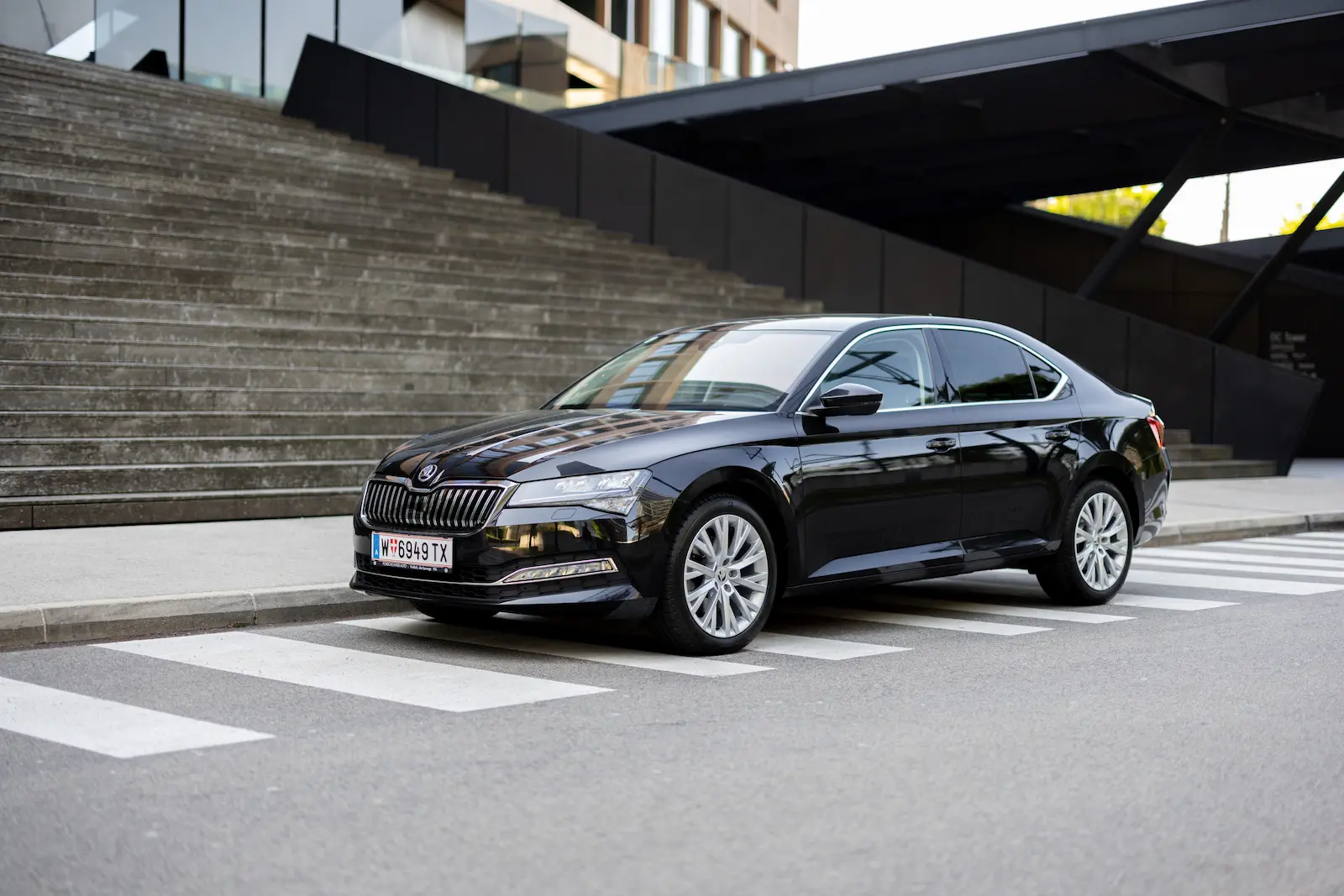 Schwarze Skoda Superb geparkt vor einer modernen Treppe mit urbanem Gebäude im Hintergrund.