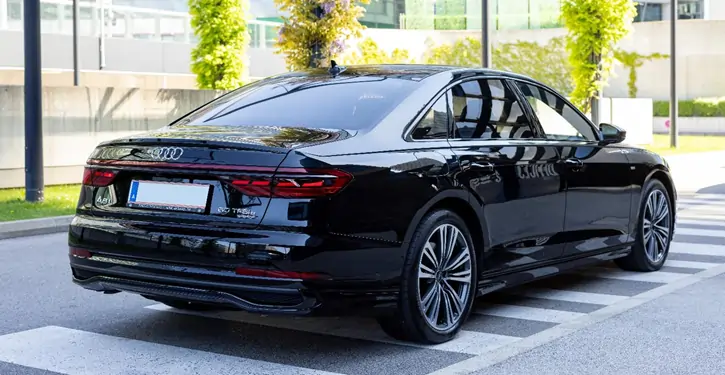 Elegante Audi A8 Long Heckansicht in Schwarz, geparkt vor einem modernen Gebäude mit grünen Pflanzen im Hintergrund.
